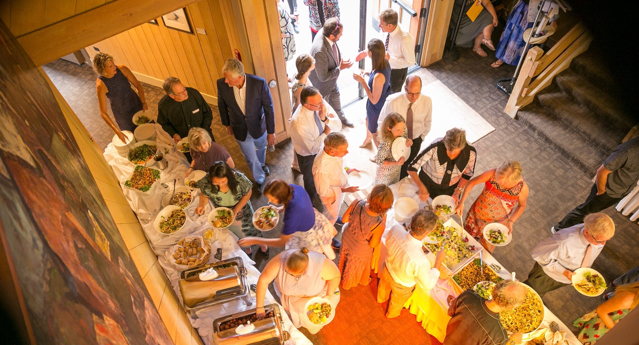 Guests serving themselves from a buffet table with salads, bread, and warm dishes during an indoor gathering at Timberline Lodge, viewed from above under warm lighting.