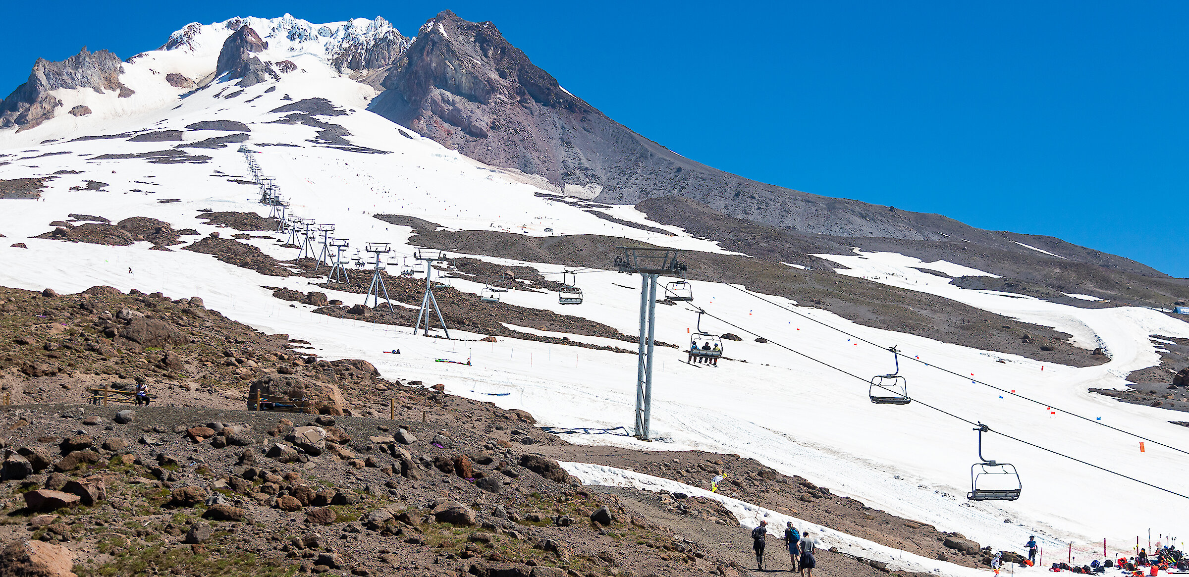 Skiers and hikers make their way up the snowy slopes of Mount Hood near Timberline Lodge, with chairlifts running alongside patches of snow and exposed rock under a clear blue summer sky.