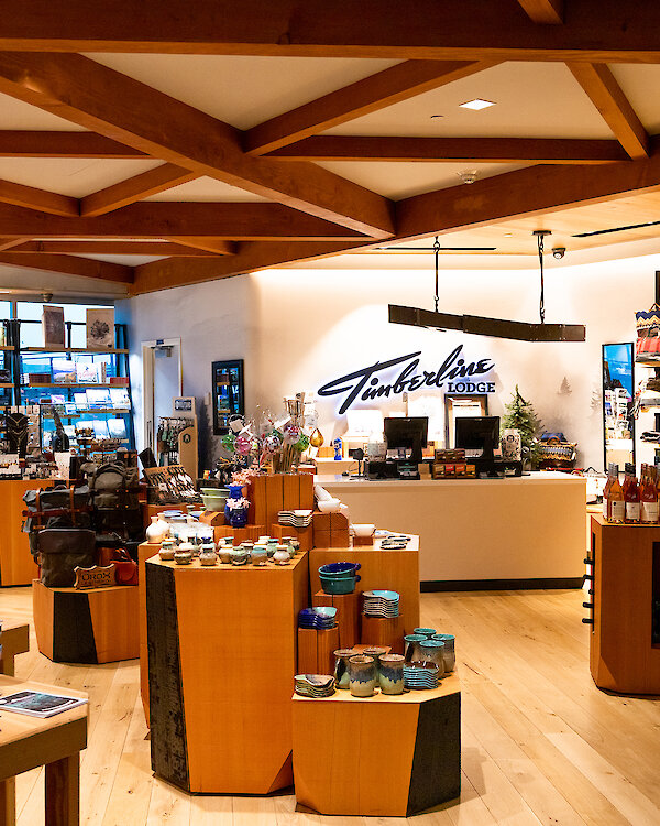 Interior of the Timberline Lodge store at Portland International Airport, featuring wooden displays with pottery, souvenirs, apparel, and Oregon-made gifts under warm lighting and a Timberline Lodge sign behind the counter.