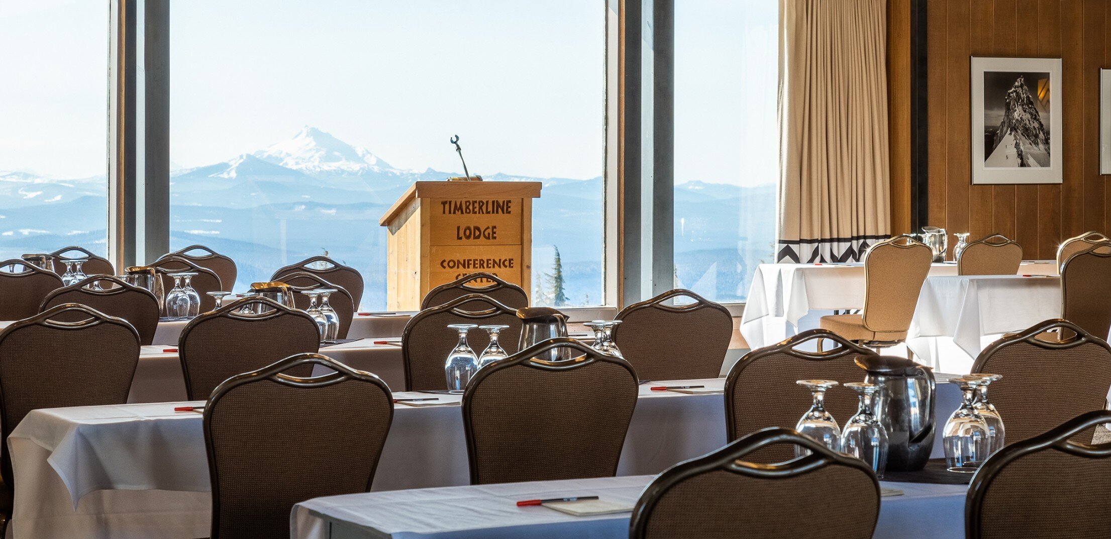 Wooden podium with "Timberline Lodge Conference Center" carved into its front pictured in front of a window featuring views of Mt. Jefferson, facing tables and chairs set up for a meeting