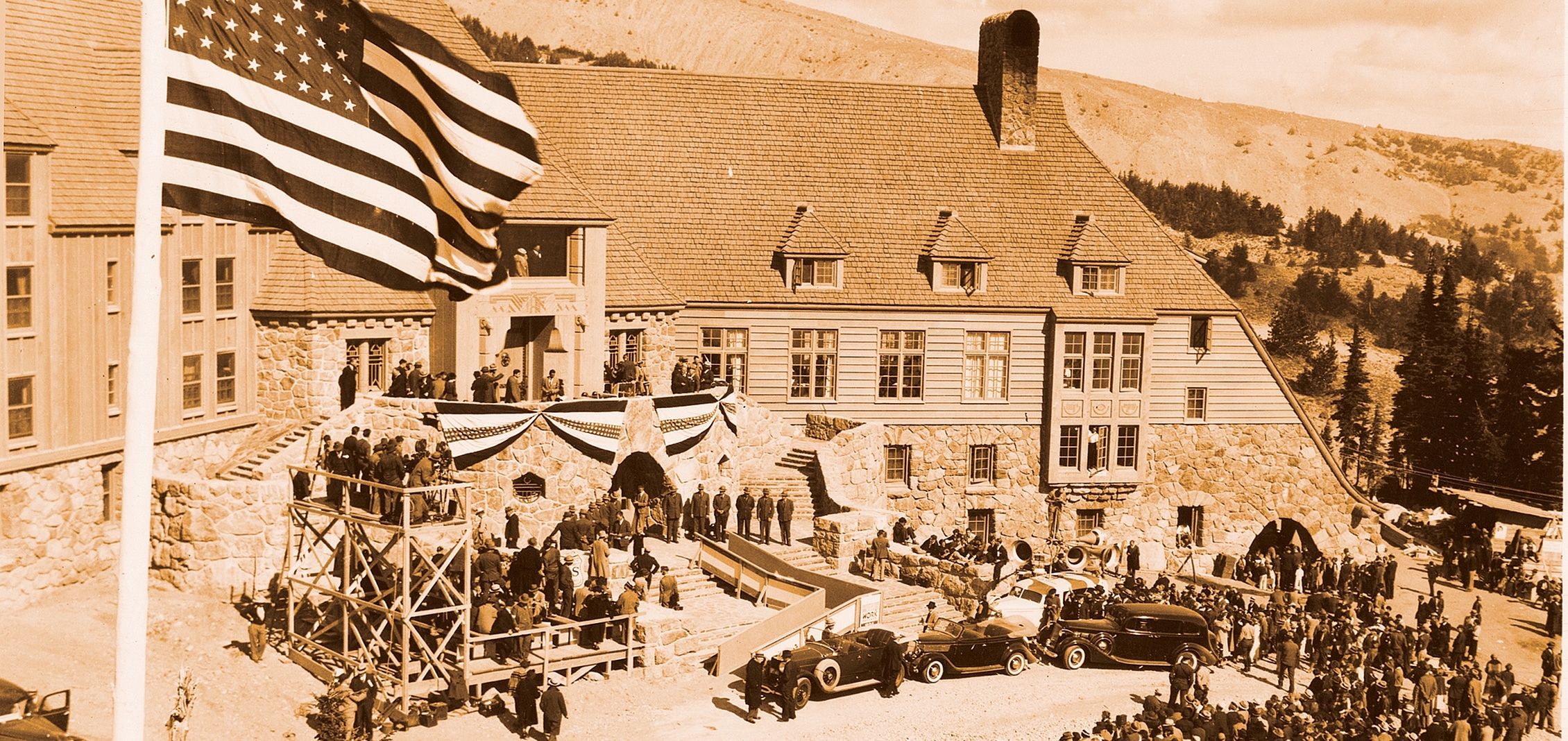 Historic photo of FDR dedication of Timberline Lodge with American flag in foreground, vintage cars in front of lodge, and crowd gathering to watch