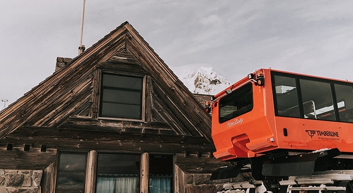 SILCOX HUT IN THE DAYTIME SNOW WITH A RED TIMBERLINE SNOWCAT IN THE FOREGROUND AND THE PEAK OF MT. HOOD BEHIND