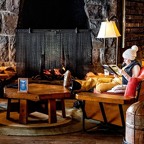 WOMAN IN WHITE BEANIE READING A BOOK ABOUT TIMBERLINE IN FRONT OF FIREPLACE