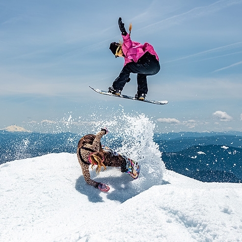 ONE FEMALE SNOWBOARDER IN PINK COAT AND BLACK PANTS JUMPING OVER FEMALE SNOWBOARDER IN LEOPARD PRINT COAT