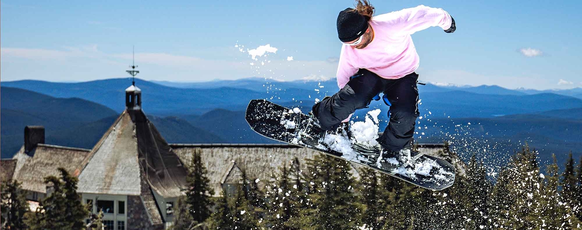 Snowboarder in a pink shirt catches air above a snowy slope in front of Timberline Lodge on a sunny day, with pine trees and distant mountains in the background.