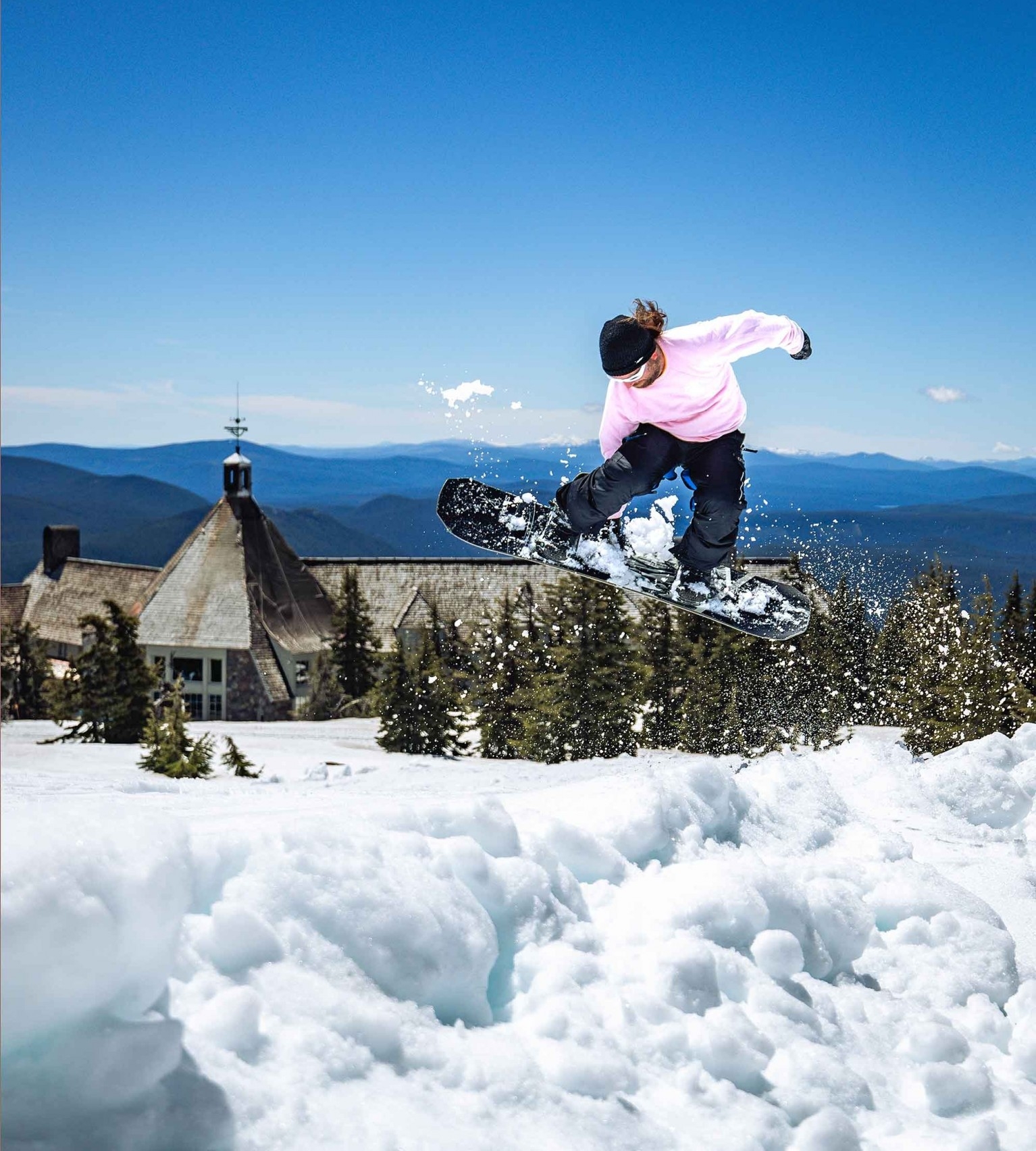 Snowboarder in a pink shirt catches air above a snowy slope in front of Timberline Lodge on a sunny day, with pine trees and distant mountains in the background.