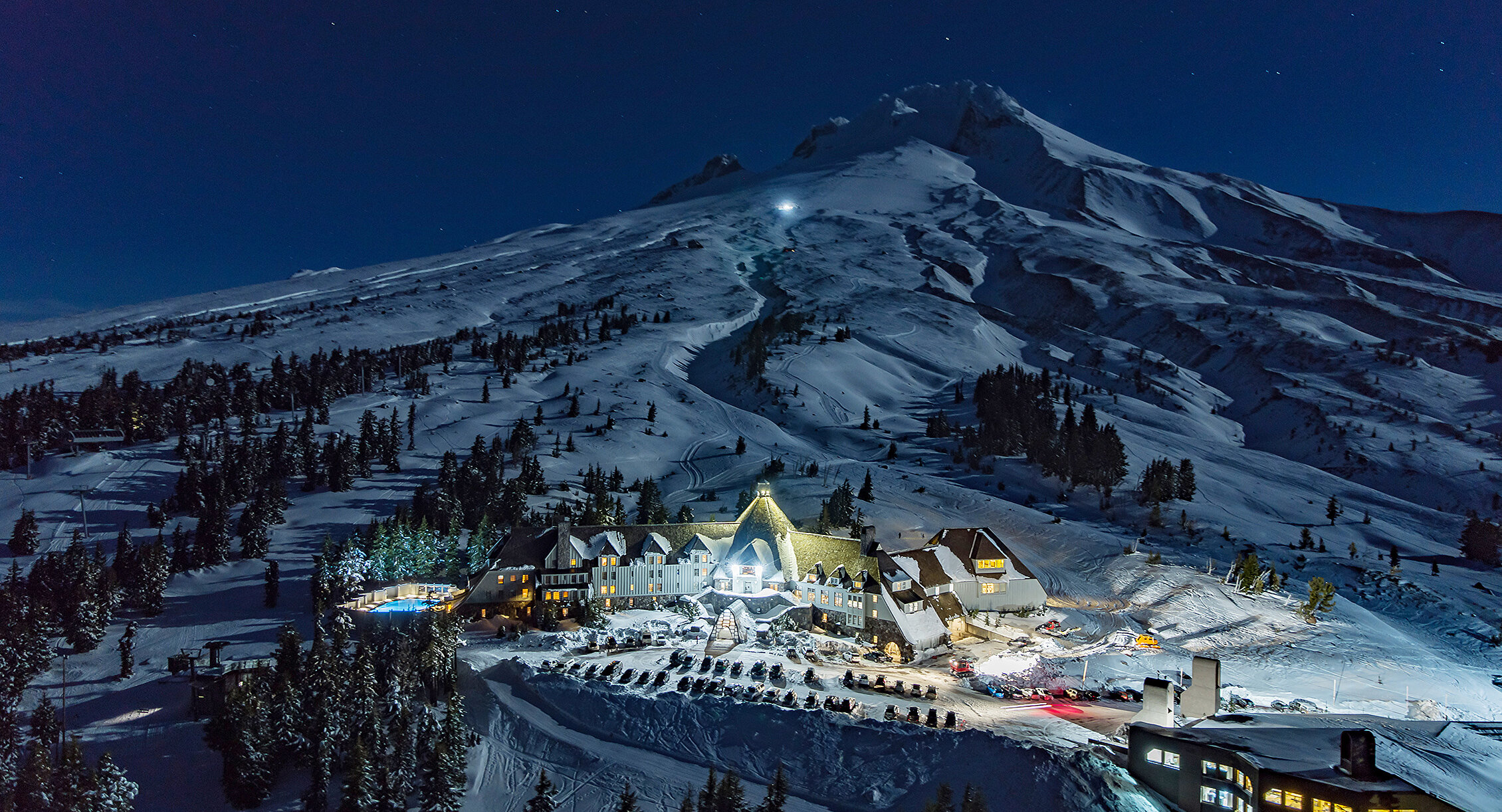 Timberline Lodge illuminated at night, surrounded by snow-covered slopes on Mount Hood under a clear, starry sky. The mountain rises steeply behind the lodge, glowing faintly in the moonlight.