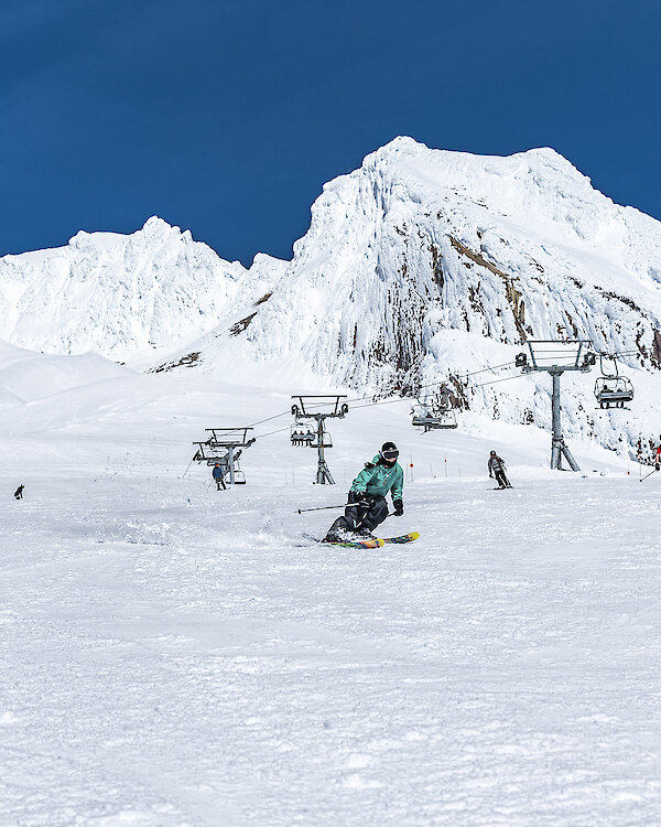 Skiers and snowboarders gliding down a snowy slope under a bright blue sky on Mount Hood at Timberline, with people in colorful winter gear, chairlifts ascending in the background, and steep, snow-covered ridges behind them.