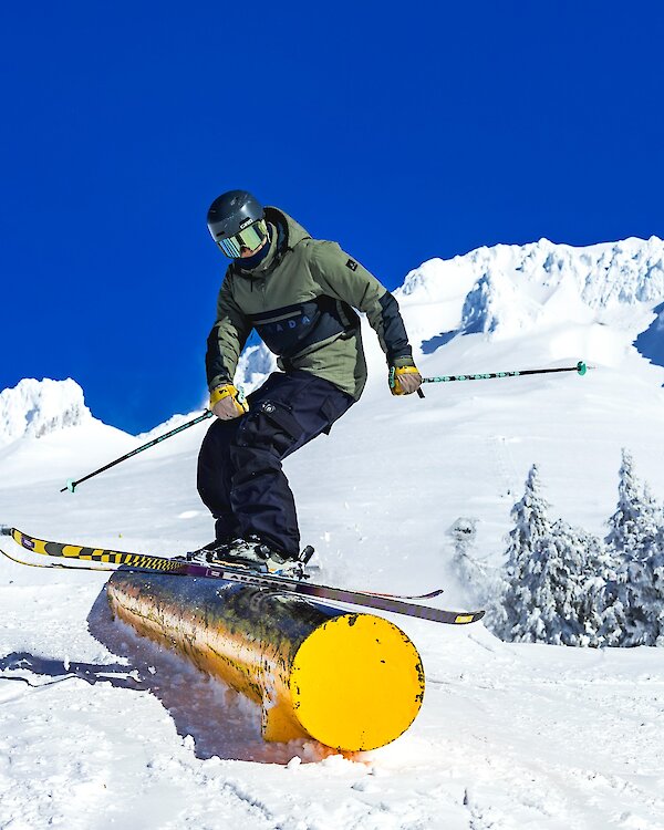 A skier in a green jacket and black helmet balances on a yellow rail feature in the snow park at Timberline Lodge on Mount Hood, with snowy slopes, trees, and a bright blue sky in the background.