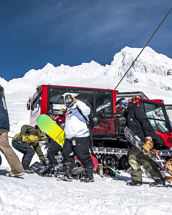 Snowboarders unload from a red snowcat on the snowy slopes at Timberline Lodge, with Mount Hood’s snow-covered peak and a bright blue sky in the background.