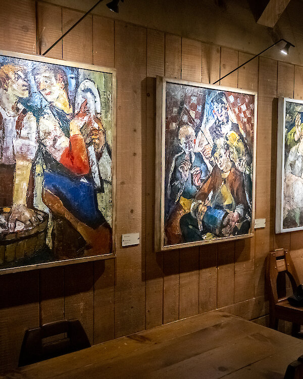 Three colorful paintings hang on a wood-paneled wall inside Timberline Lodge, depicting stylized figures in expressive poses under warm lighting above rustic wooden tables and chairs.
