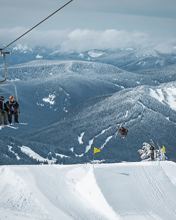 Skier performing a jump near a chairlift at Timberline Lodge with snow-covered mountains and trees in the background.
